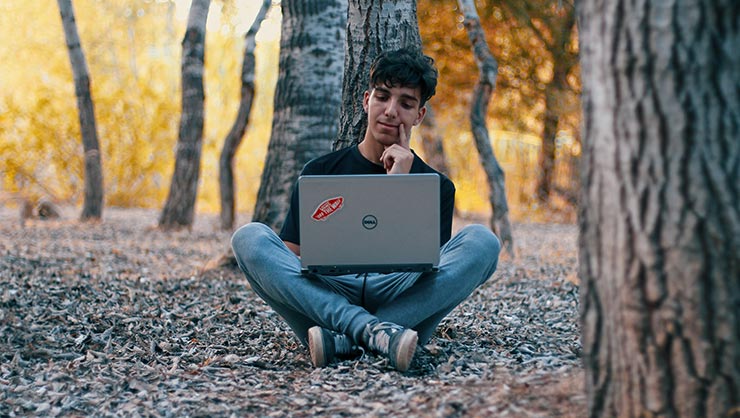 A boy sits in the woods with a laptop